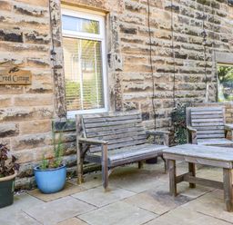 An outdoor seating area with wooden benches and table next to a stone wall with a window and potted plants at Creel Cottage Blackwood Street in Amble