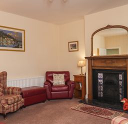 A sitting room with armchairs a fireplace a wooden side table with a lamp and framed pictures on the walls at Creel Cottage Blackwood Street in Amble