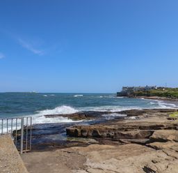 A rocky beach with waves crashing a metal railing a row of houses on a grassy coastline and an island with a lighthouse in the sea at Creel Cottage Blackwood Street in Amble