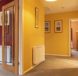 A hallway with yellow walls, carpeted floor, framed pictures on walls, and a wooden door with glass panels at Our Story Cottage Longhoughton