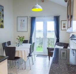 A dining area with a round table and chairs next to glass doors with curtains and a small armchair at Our Story Cottage in Longhoughton