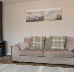 A living room with a beige sofa with plaid cushions a wood burning stove and a painting above the sofa at Southfield Lodge in Seahouses