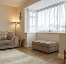 A living room with a sofa a floor lamp an ottoman a television and a bay window with blinds at Southfield Lodge in Seahouses