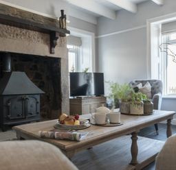 A living room with a wood burning stove a wooden coffee table with cups and pastries and a grey armchair by a window at Creel Cottage Craster