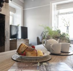 A table with a plate of scones and strawberries and a teapot with cups in a living room with a television and plants at Creel Cottage Craster