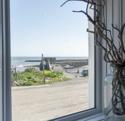 A view of a harbor and ocean through a window with a glass jar holding twigs on the windowsill at Creel Cottage Craster
