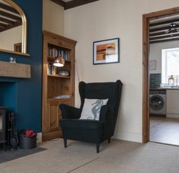 A living room with a black armchair, wooden bookshelf, fireplace, and a view into a kitchen with a washing machine at Kittiwake Cottage Seahouses