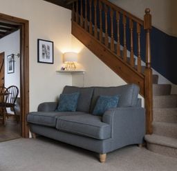 A living room with a gray sofa and cushions next to wooden stairs and a dining room with wooden chairs in the background at Kittiwake Cottage Seahouses