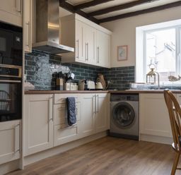 A kitchen with white cabinets a washing machine wooden dining chairs and a window at Kittiwake Cottage Seahouses
