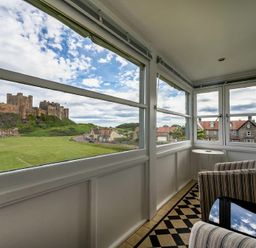 An enclosed balcony with two striped chairs a small round table and large windows overlooking a castle and houses at Keepers View in Bamburgh
