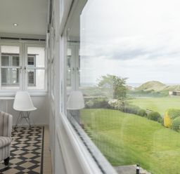 A narrow room with a striped armchair a white chair and windows overlooking a garden at Keeper's View in Bamburgh