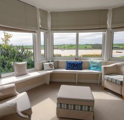 A seating area with armchairs a bench with cushions an ottoman and large windows overlooking water and greenery at The Beach House Alnmouth