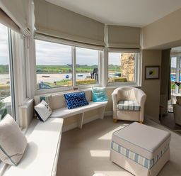 A seating area with window seats, cushions, an armchair, and an ottoman overlooking a waterfront at The Beach House, Alnmouth