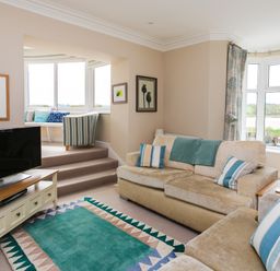 A living room with beige sofas and striped cushions a teal rug a television on a wooden stand and a small raised seating area at The Beach House Alnmouth