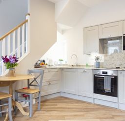 A kitchen with light gray cabinets wooden floor a small wooden table with two chairs and flowers in a vase under a staircase at Wisteria Cottage in Warkworth