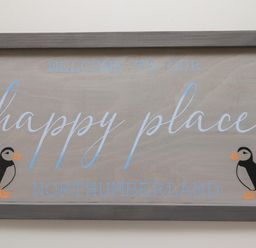 A wooden sign with the text welcome to our happy place northumberland and two puffin illustrations