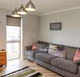 A living room with a gray sofa with multiple cushions a wooden side table and a wall clock at Puffin View in Seahouses