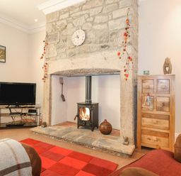 A living room with a stone fireplace and wood stove a television on a metal stand and a wooden chest of drawers at Willow Cottage in Lesbury