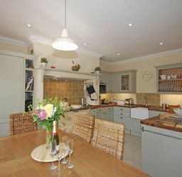 A kitchen with wooden table and woven chairs and cabinets with a clock on the wall at Grange Cottage in Alnmouth