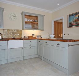A kitchen with grey cabinets wooden countertops a white sink clock on the wall and a Bamburgh poster at Grange Cottage in Alnmouth