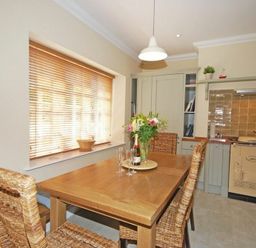 A dining area with a wooden table and wicker chairs next to a window with blinds and a kitchen with beige cabinets and an Aga cooker at Grange Cottage in Alnmouth