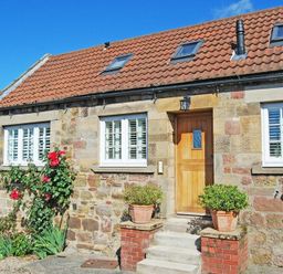 Front exterior of a stone cottage with a wooden door red flowers and potted plants on steps at Martins Cottage in Alnmouth