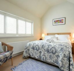 A bedroom with a bed with patterned bedding two lamps on wooden bedside tables a wicker chair with cushions and a window with blinds at Martins Cottage in Alnmouth