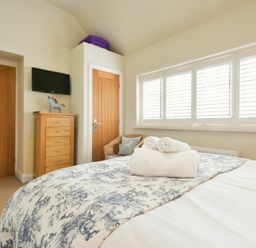 A bedroom with a bed covered in a patterned blanket and rolled towels windows with shutters a wooden chest of drawers a wall-mounted television a wicker chair and two wooden doors at Martins Cottage in Alnmouth