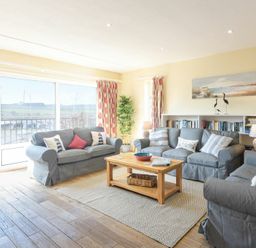 A living room with three gray sofas a wooden coffee table floor lamp bookshelf and large windows with curtains at The Boathouse Alnmouth