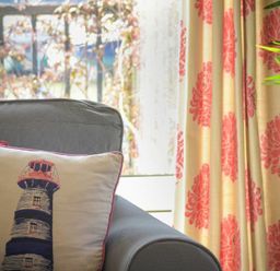 A cushion with a lighthouse design on a grey armchair next to floral patterned curtains and a plant at The Boathouse (Alnmouth) in Alnmouth