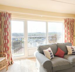 A living room with a gray sofa and patterned curtains by large windows overlooking a marina at The Boathouse (Alnmouth) in Alnmouth