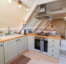 A kitchen with grey cabinets wood countertops a stove and sink with a living area with a television and sofa in the background at Coach House Alnmouth