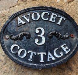 A black oval house number plaque with white letters and the number 3 on a stone wall at Avocet Cottage Bamburgh