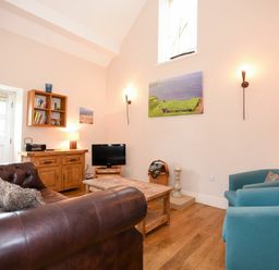 A living room with a brown leather sofa two blue armchairs a wooden coffee table a wooden cabinet with shelves a small television and wall art at High Tide in Newton-by-the-Sea