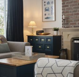 A living room with a sofa a wooden coffee table a dark dresser with a lamp and a fireplace at Sea Croft in Beadnell