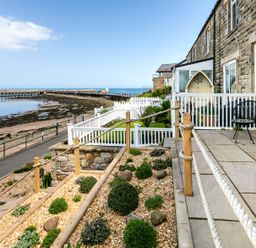 A stone building with a patio and garden overlooking a pier and the sea at Old Coastguard Cottages in Amble
