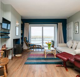 A living room with beige sofa red leather chair coffee table and sliding glass door overlooking water at Old Coastguard Cottages in Amble