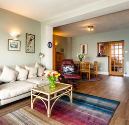 A living room with a beige sofa, a glass coffee table, a colorful rug, a red armchair, a wooden dining table with chairs, and wall art at Old Coastguard Cottages in Amble