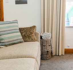 A beige sofa with two cushions next to a woven basket holding folded blankets by a window at Armstrong Cottages No30 in Bamburgh