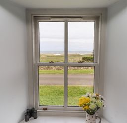 A window with a view of grass road and ocean with binoculars and a vase of flowers on the windowsill at Beachward in Embleton