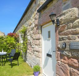 A stone exterior wall with a white door and a black outdoor light next to a garden table and chairs at Lavender Cottage Village Farm in Seahouses