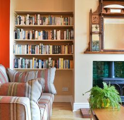 A living room with a striped sofa a bookshelf filled with books a wood stove with green tiled surround and a wooden mirror above at Estuary View in Alnmouth