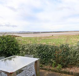 An outdoor patio with a glass table and four chairs next to a hedge with a grassy field and water in the background at Kittling Cottage in Bamburgh