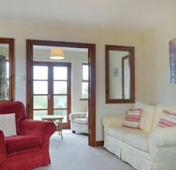 A living room with a red armchair a white sofa with striped pillows wooden framed mirrors and a view into a room with glass doors at Smithy Court in Craster