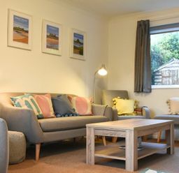 A living room with a sofa, armchairs, cushions, a coffee table, two lamps, framed pictures on the wall, and a window showing a garden shed at Barmoor in Embleton