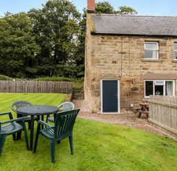 A garden with plastic chairs and a round table beside a stone house with wooden fencing at Bilton Farm Cottage No1 in Alnmouth