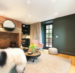 A living room with a brick fireplace a round mirror a pink armchair and a coffee table with flowers at The Bolthole in Eglingham