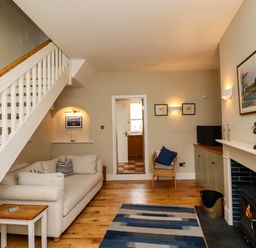 A living room with a sofa under stairs a wood burning stove and a doorway to a kitchen at Strandline in Alnmouth