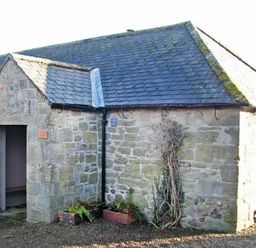 An exterior view of a stone building with a slate roof and an open doorway with plants outside at Swallow Cottage nr Powburn in Powburn