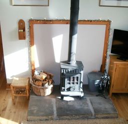 A wood-burning stove with a basket of firewood and a metal ash container next to a wooden cabinet and stool at Swallow Cottage nr Powburn in Powburn